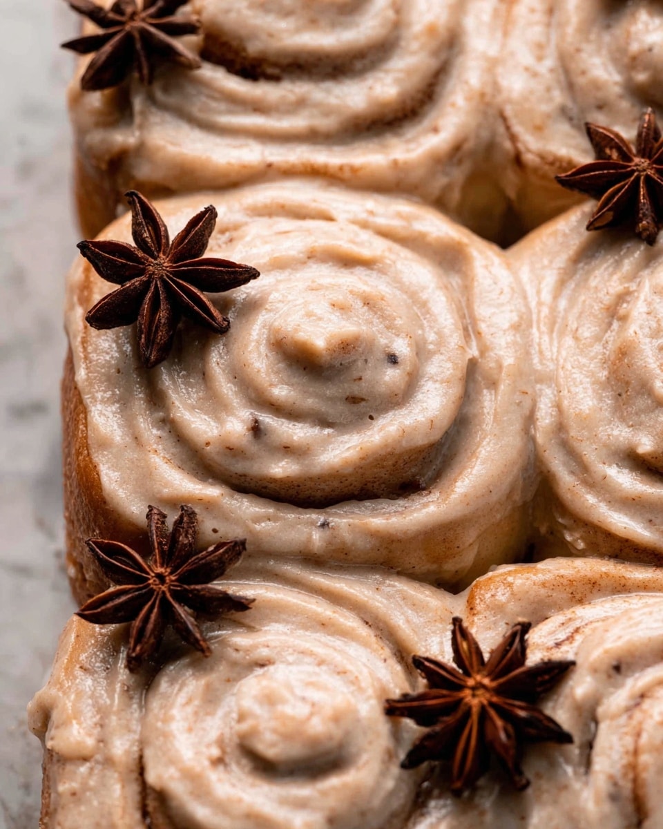 A close-up view of a group of soft cinnamon rolls covered in a creamy, light brown cinnamon frosting that looks smooth and thick, with swirled textures visible on each roll. The rolls are tightly packed together, and there are dark brown star anise pieces placed on top as decoration, adding contrast. The background features a white marbled texture. photo taken with an iphone --ar 4:5 --v 7