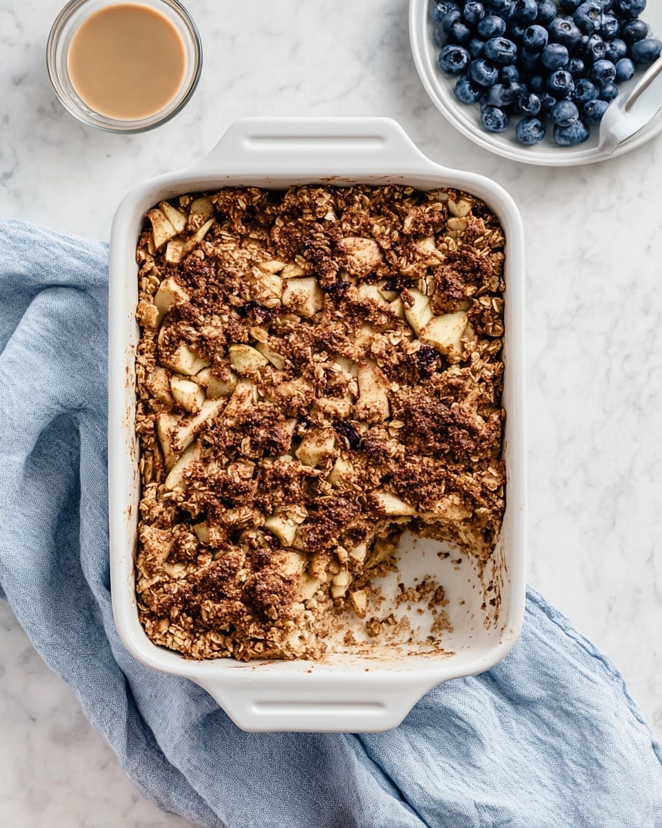 The image shows a white rectangular baking dish filled with a baked oat and fruit mixture. The top layer is uneven and golden brown with visible chunks of apple pieces and oats spread throughout, creating a rough texture with some darker spots from baking. A noticeable scoop is missing from one corner, revealing a moist, dense inside layer of the oat mixture with softer apple bits embedded. The baking dish rests on a soft, crumpled blue and white cloth against a white marbled background. To the right, there is a white plate with fresh blueberries, and to the left bottom corner, a small clear glass bowl containing a light brown creamy sauce. Photo taken with an iphone --ar 4:5 --v 7