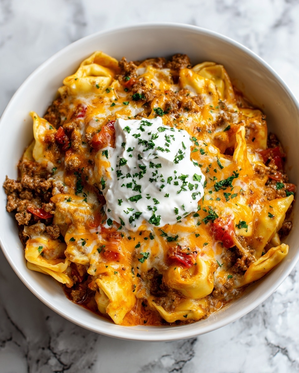 A white bowl filled with several layers of tortellini pasta mixed with ground meat, showing light yellow pasta and brown meat with small bits of red tomato. On top, a generous layer of melted cheddar and mozzarella cheese covers the pasta, creating an orange and white melted texture. A dollop of white sour cream sits in the center, sprinkled with small pieces of chopped green herbs. The bowl is placed on a white marbled surface, focusing closely on the warm, cheesy dish. photo taken with an iphone --ar 4:5 --v 7
