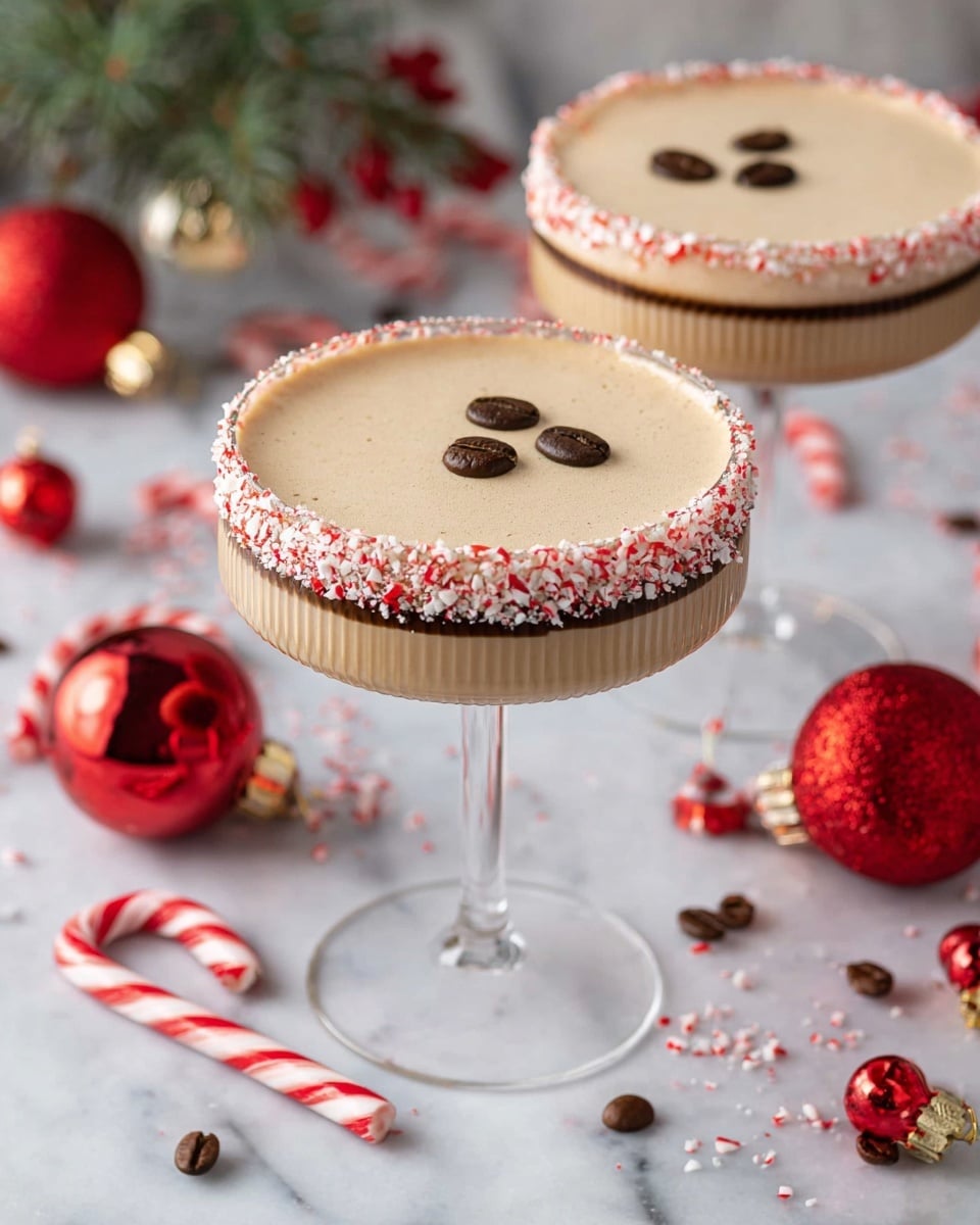Two creamy beige cocktails sit in tall, clear stemmed glasses with ridged white bowls. Both glasses have rims coated in crushed red and white peppermint candy and a thin dark chocolate layer beneath it. Each drink's smooth top layer is decorated with three dark brown coffee beans arranged in a triangle. The glasses are placed on a white marbled surface scattered with red shiny Christmas ornaments, whole coffee beans, and red-and-white striped candy canes, creating a festive holiday feel. Photo taken with an iphone --ar 4:5 --v 7
