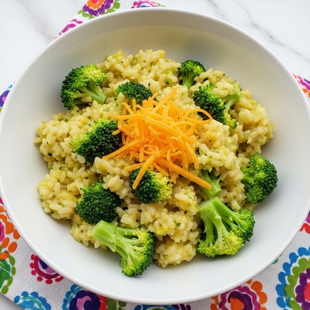 The image shows a close-up of a bowl filled with creamy broccoli risotto. The risotto has a thick, slightly glossy texture with small, soft grains of rice mixed evenly with small pieces of bright green broccoli. The creamy sauce coats every grain, giving it a light yellow color mixed with green specks from the broccoli. The bowl is white, and in the background, there is a blurry olive green bowl with white spots. The surface beneath is a white marbled texture with a colorful cloth partially visible near the bottom right corner. photo taken with an iphone --ar 4:5 --v 7