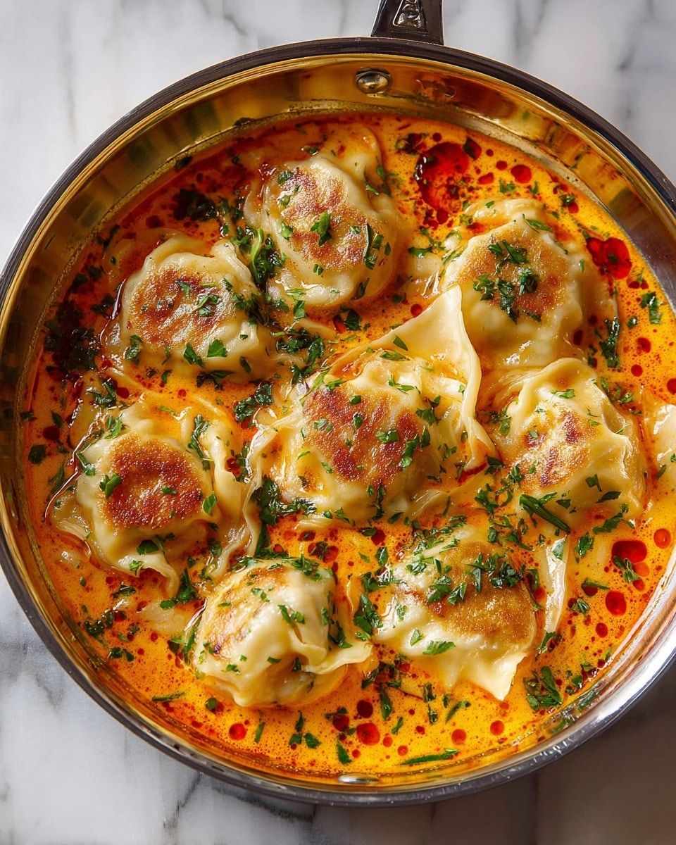 A round silver pan filled with seven dumplings floating in a bright orange creamy sauce with red oil spots, each dumpling has a light golden-brown sear on top and soft, slightly translucent edges. The dish is topped with finely chopped green herbs scattered evenly across the dumplings and sauce. The pan is set on a white marbled surface. photo taken with an iphone --ar 4:5 --v 7