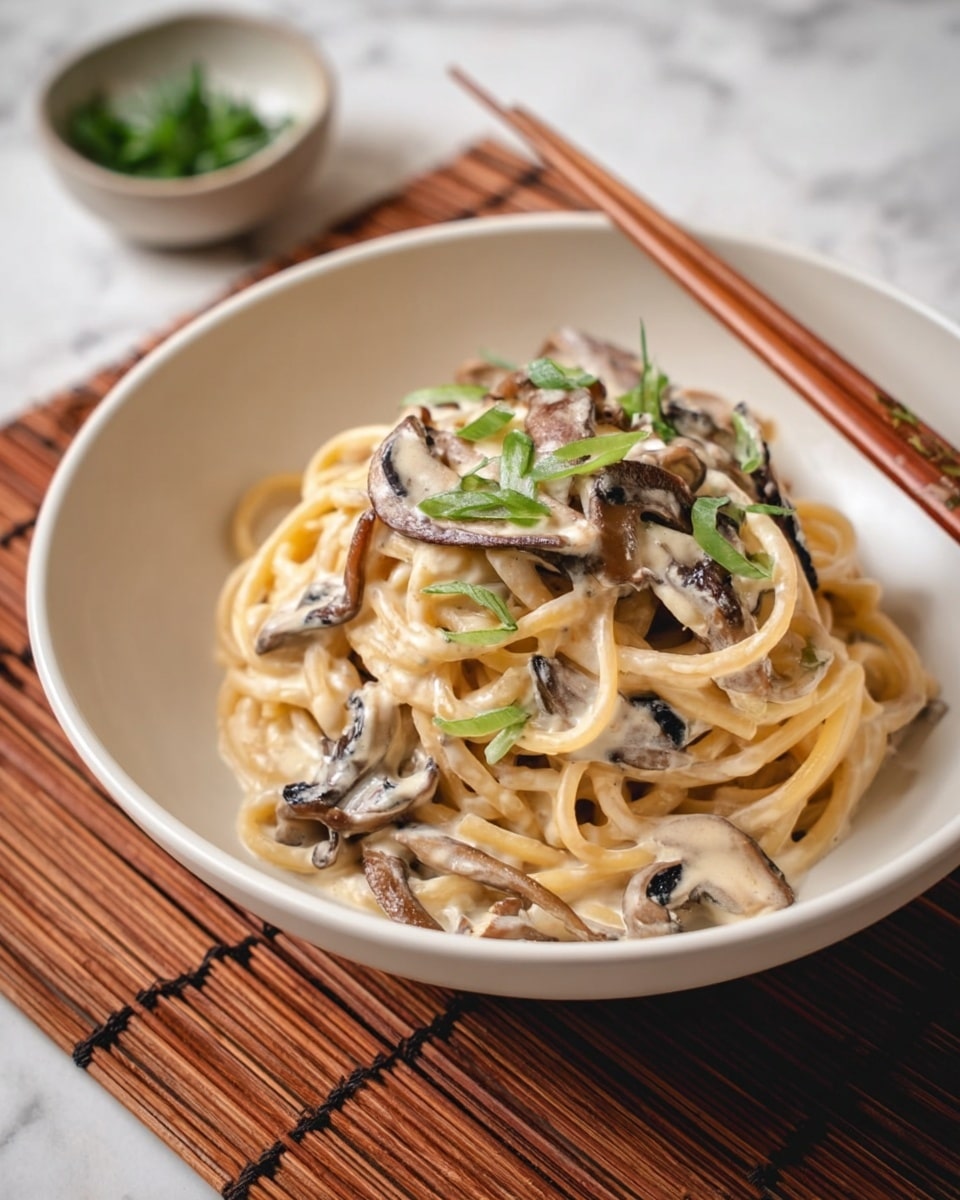 A white bowl filled with a small mound of thick spaghetti noodles coated in a light creamy sauce, mixed with thin, dark brown and white striped mushroom slices spread throughout the pasta. The dish is garnished with scattered small green onion rings on top and around the noodles. The bowl rests on a bamboo mat with a pair of wooden chopsticks placed nearby. In the background, there is a white bowl with green herbs, all set against a white marbled surface. photo taken with an iphone --ar 4:5 --v 7