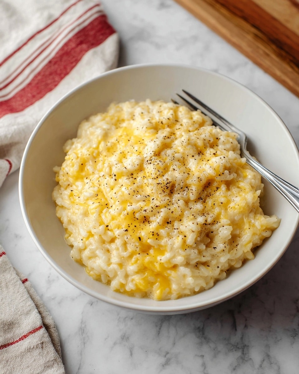 A white bowl filled with creamy, cheesy risotto, showing one main layer of soft, cooked rice mixed with melted yellow cheese that creates a rich, smooth texture; the dish is sprinkled lightly with black pepper for a touch of detail on top. The bowl sits on a white marbled surface next to a silver fork, with a wooden cutting board and a red-striped beige cloth in the soft-focus background. photo taken with an iphone --ar 4:5 --v 7