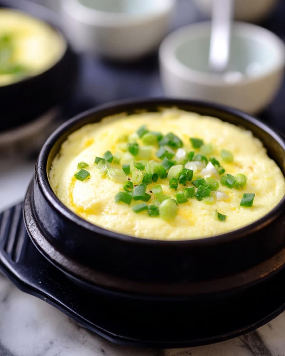 The image shows a black round bowl filled with a thick, fluffy layer of pale yellow steamed egg custard. On top of the smooth egg layer is a scattering of chopped bright green scallions, adding texture and color. The bowl rests on a stand, all set against a white marbled surface. In the blurry background, there are white bowls and a spoon. photo taken with an iphone --ar 4:5 --v 7