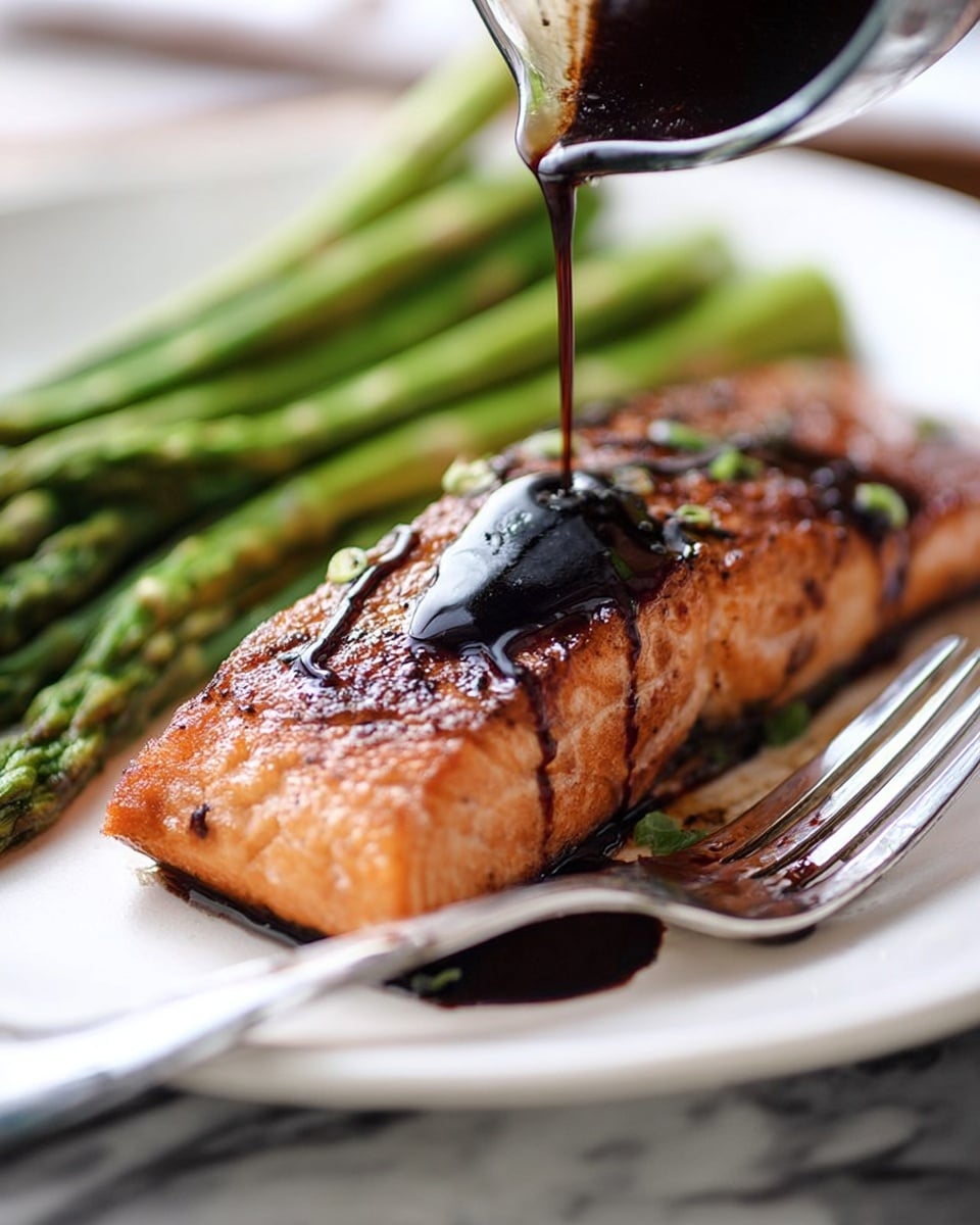 A piece of cooked salmon with a crispy brown top layer and moist orange-pink inside sits on a white plate. Dark, shiny balsamic glaze is being poured over the salmon, adding a glossy black layer on top. To the right of the salmon, there is a bundle of bright green cooked asparagus with a smooth texture. A shiny silver fork rests on the plate’s edge near the asparagus. The plate is set on a white marbled surface, and the overall look is warm and inviting. photo taken with an iphone --ar 4:5 --v 7