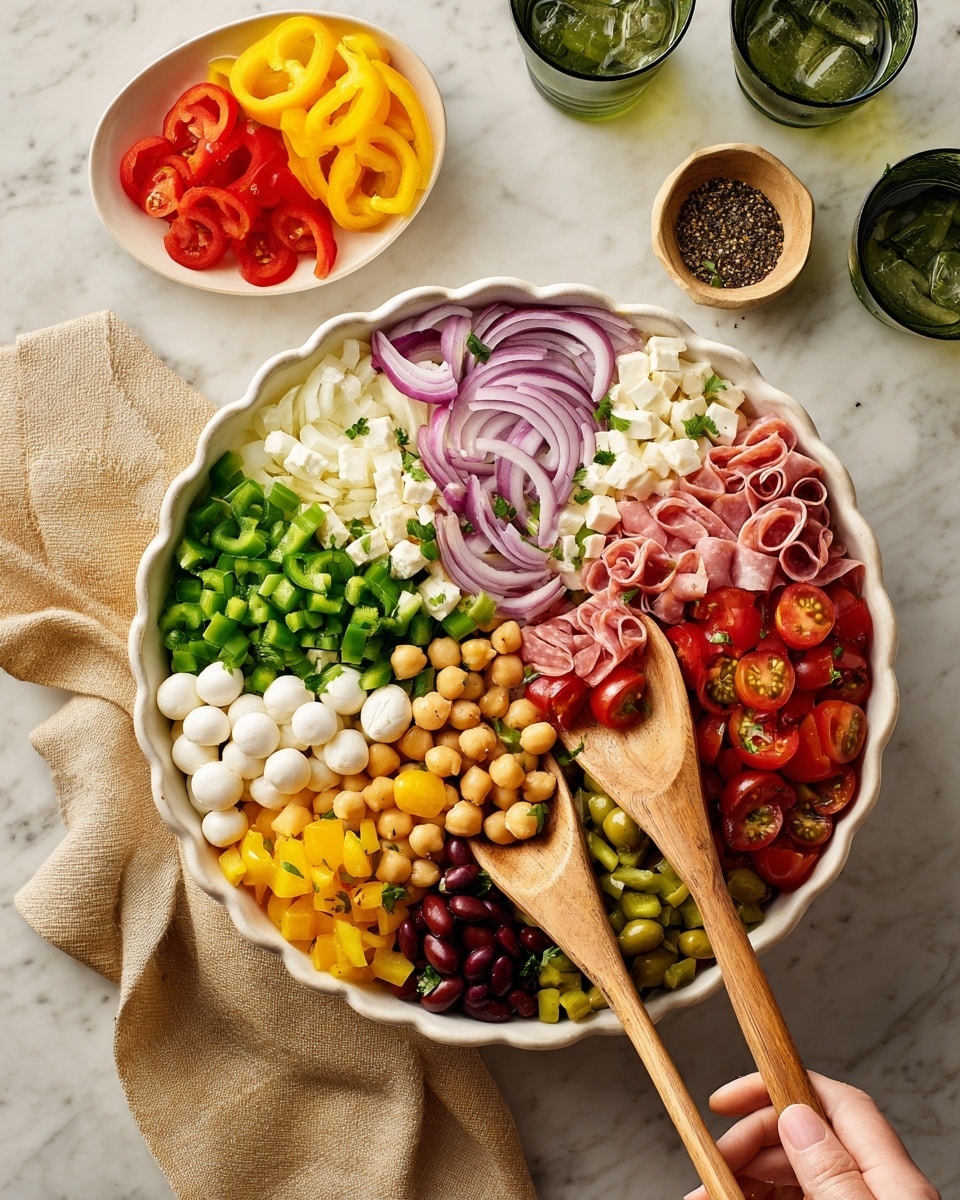 A large white scalloped bowl filled with a vibrant mix of salad ingredients is being tossed with two wooden spoons held by a woman's hand at the bottom right. The salad has distinct layers including sliced red onions with a smooth texture, bright green chopped bell peppers, red kidney beans with a shiny surface, beige chickpeas, small white mozzarella balls with a soft look, chopped cherry tomatoes with a glistening cut surface, and thin strips of pink and red deli meats scattered across the top. The bowl sits on a white marbled surface, with a beige cloth to the bottom left, a small white bowl of sliced yellow and red peppers above, and a small white dish of black pepper to the right. Two green and black glasses with ice rest near the top left. Photo taken with an iphone --ar 4:5 --v 7