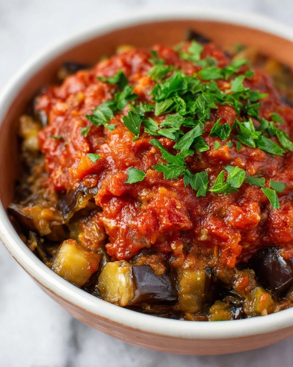 A close-up of a dish served in a white bowl showing two visible layers: the bottom layer is chunky pieces of roasted eggplant with a soft, slightly glistening texture in brown and light yellow tones, and the top layer is a thick, rich red tomato sauce with small diced pieces and a slightly chunky texture, garnished with bright green parsley leaves scattered on top. The bowl is placed on a white marbled surface. photo taken with an iphone --ar 4:5 --v 7