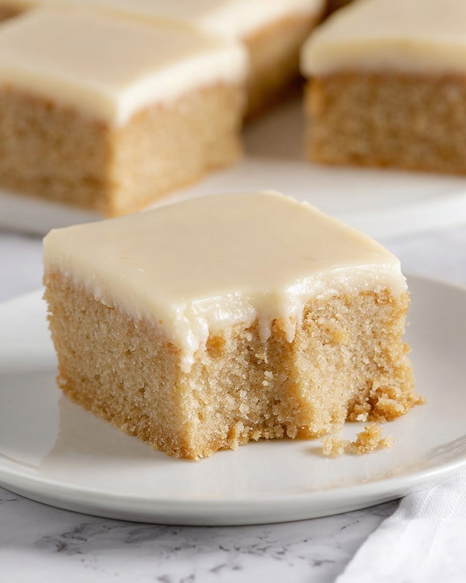A square piece of cake with two layers sits on a white plate over a white marbled surface. The bottom layer is thick, light brown, and crumbly with a soft texture. The top layer is a smooth, pale cream color, slightly glossy, appearing as a thin frosting spread evenly across the cake. One corner of the cake has a bite taken out, showing the contrast between the two layers clearly, and some crumbs are scattered near the plate's edge. In the background, another piece of the same cake is slightly out of focus. photo taken with an iphone --ar 4:5 --v 7
