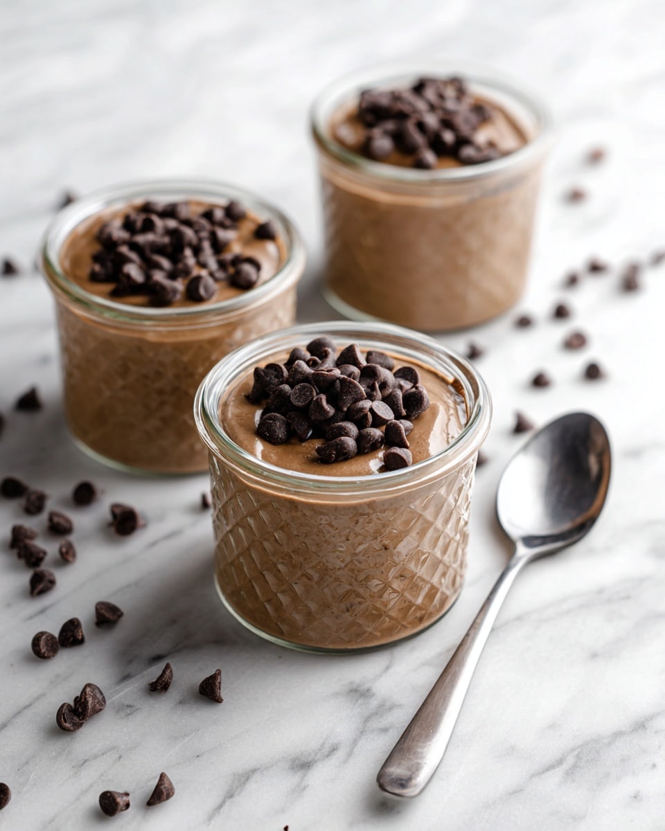 Three clear glass jars filled with smooth, light brown chocolate pudding, each topped generously with a pile of dark chocolate chips. The jars are placed on a white marbled surface, with some chocolate chips scattered around. To the right of the jars, there is a shiny metal spoon resting on the surface. The texture of the pudding looks creamy, and the glass jars have a subtle pattern that catches light softly. Photo taken with an iphone --ar 4:5 --v 7