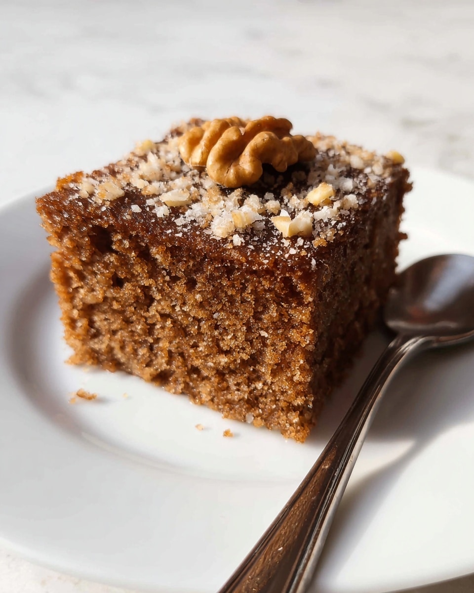The image shows a single square piece of moist brown cake with a coarse texture, topped with a scattering of light-colored crumbs and a whole walnut placed neatly in the center on top. The cake is placed on a simple white plate with a silver spoon lying next to it. The background is a white marbled surface. photo taken with an iphone --ar 4:5 --v 7