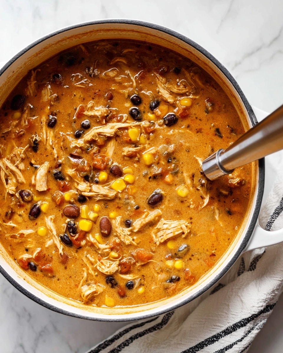 A close-up view of a white pot filled with a thick, creamy soup that has a rich orange-brown base mixed with shredded pieces of chicken, small black beans, bright yellow corn kernels, and visible bits of cooked onions and tomatoes, creating a colorful, chunky texture. A metal ladle with a wooden handle rests inside the pot on the right side, partially submerged in the soup. The pot is sitting on a white marbled surface with a folded white cloth with black stripes peeking from underneath on the bottom right. photo taken with an iphone --ar 4:5 --v 7
