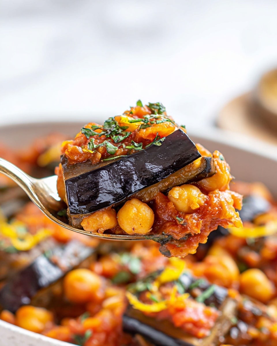 A close-up image shows a spoon holding a layered piece of cooked eggplant with a shiny dark purple-black skin on top. Below the eggplant layer is a thick layer of orange chickpeas mixed with soft, diced onions and red tomato sauce, with small pieces of green herbs sprinkled around. The background shows more chunks of the same dish in a white bowl, with the same mix of orange chickpeas, red tomato, and dark eggplant pieces, garnished with green herb leaves and small yellow lemon zest pieces. The whole scene sits on a white marbled surface. photo taken with an iphone --ar 4:5 --v 7