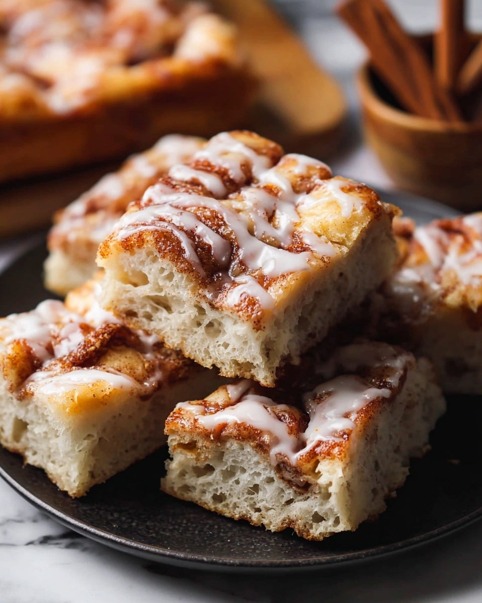 The image shows several square pieces of cinnamon roll focaccia bread stacked on a dark plate. Each piece has a thick, soft-looking dough base with visible air holes, topped with a golden-brown layer of swirled cinnamon and sugar. A glossy, white icing is drizzled unevenly on top, melting slightly into the textured cinnamon layer. The background includes a blurred bowl and some cinnamon sticks resting on a white marbled surface. The focus captures the soft texture and warm colors of the bread closely. photo taken with an iphone --ar 4:5 --v 7