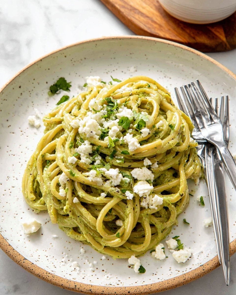 A plate with one main layer of thick spaghetti pasta coated evenly in a creamy light green sauce, topped with small crumbles of white cheese and scattered pieces of fresh green herbs, sitting on a white marbled surface, with two stainless steel forks placed to the right of the plate. The plate is white with small brown speckles and has a simple round shape. Photo taken with an iphone --ar 4:5 --v 7