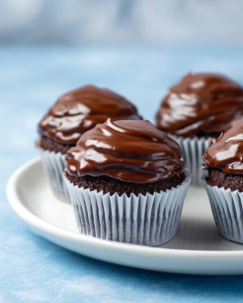 The image shows four chocolate cupcakes on a white plate, placed on a blue surface. Each cupcake is in a white paper liner with a dark cake base. The top layer is a thick, glossy chocolate frosting with a smooth, slightly swirled texture that catches the light with shiny reflections. The cupcakes have no other visible decorations and sit close together on the plate. The background is softly blurred in a light blue tone. photo taken with an iphone --ar 4:5 --v 7
