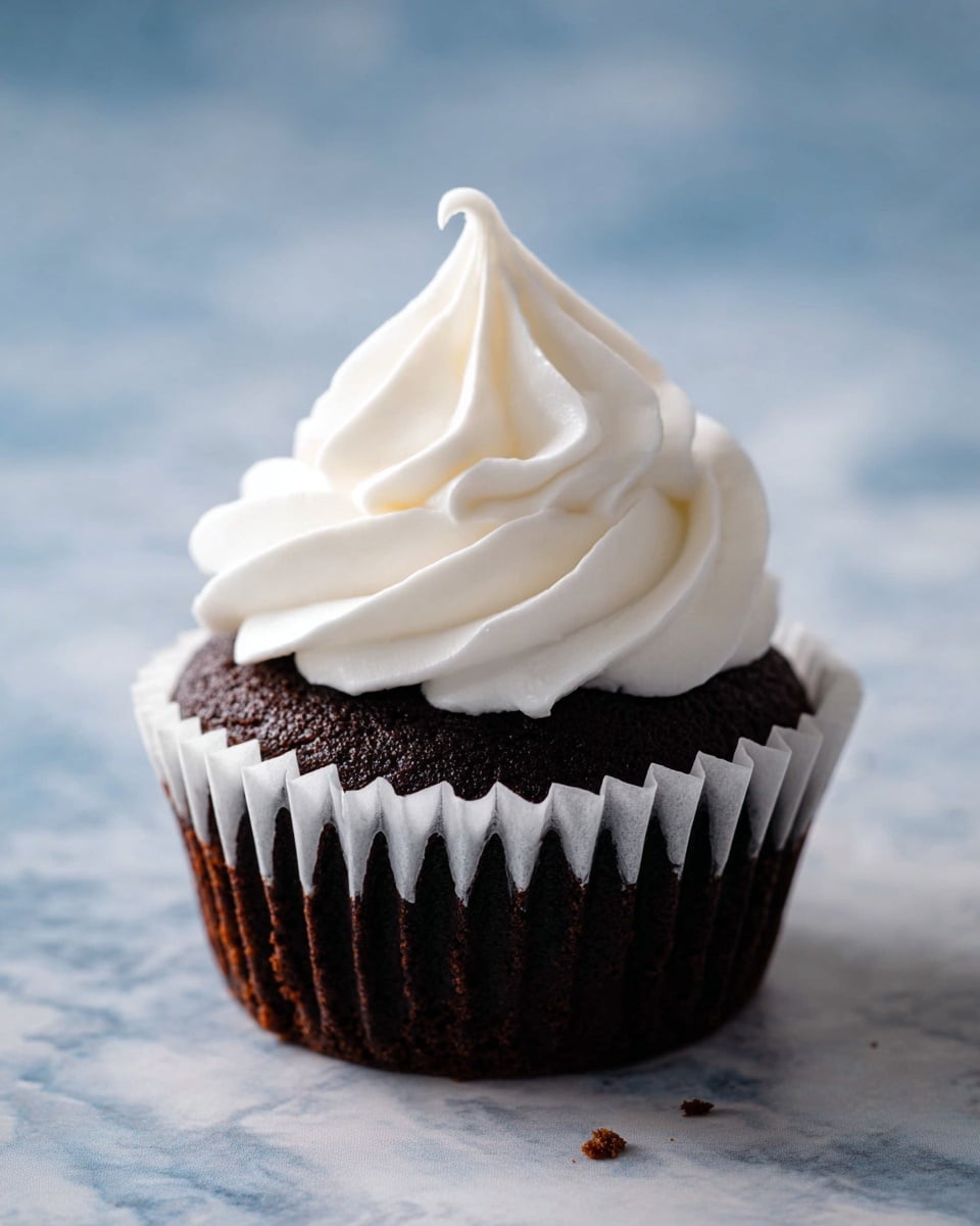 A close-up image shows a single dark brown chocolate cupcake mostly unwrapped with a white paper liner pulled down around its base. On top of the cupcake is a tall swirl of smooth, white whipped frosting that tapers to a small peak with a loop at the top. The cupcake sits on a surface with a white marbled texture in the background that is softly out of focus. The small crumb near the base adds detail to the scene. Photo taken with an iphone --ar 4:5 --v 7