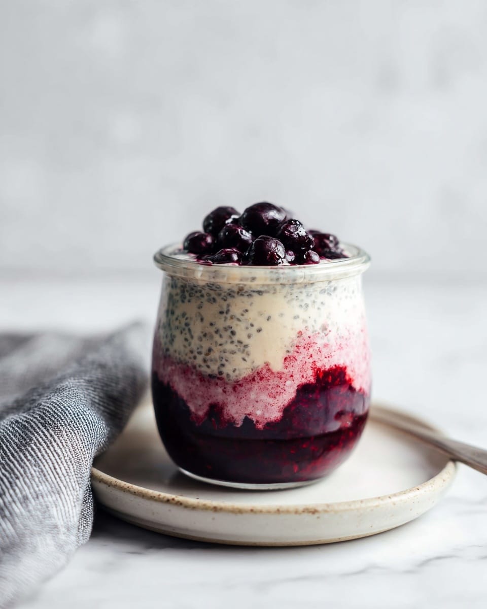 A clear glass jar shows three distinct layers: the bottom layer is a deep purple berry compote with a smooth, slightly thick texture; the middle layer features a creamy, light pink mixture with visible chia seeds giving a speckled effect; the top layer is a creamy white with chia seeds, slightly thicker in texture; on top sits a small pile of whole, dark berries with a glossy finish. The jar is placed on a round white plate with a smooth edge, set on a white marbled surface, with a folded grey and white striped cloth nearby. photo taken with an iphone --ar 4:5 --v 7