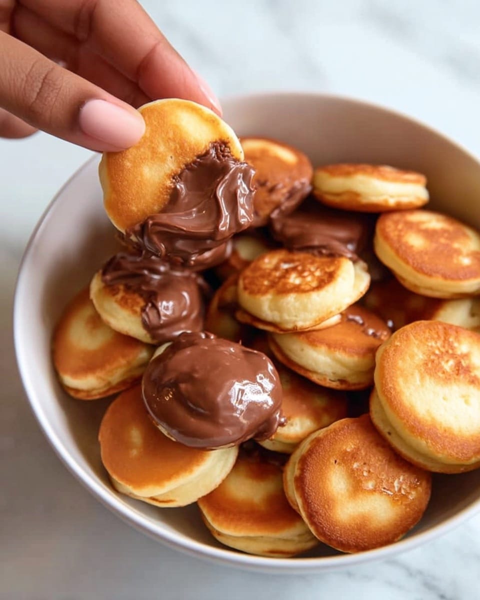 A white bowl filled with many small round golden brown mini pancakes with crispy edges and soft centers, one mini pancake held above the bowl by a woman's hand, topped with a thick layer of shiny, smooth, melted chocolate spread, some mini pancakes in the bowl also have chocolate spread visible, all placed on a white marbled surface. photo taken with an iphone --ar 4:5 --v 7