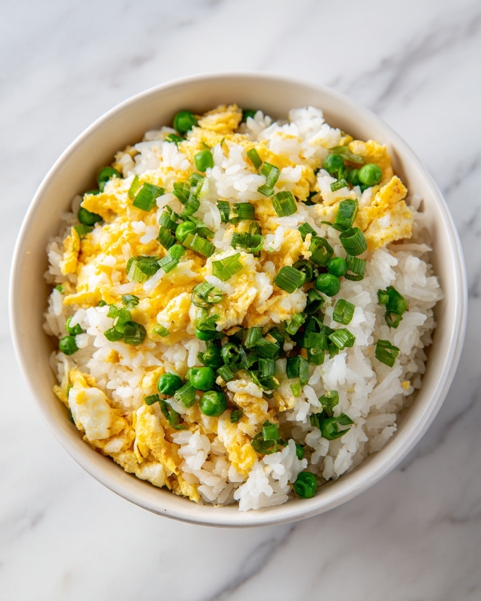 A bowl filled with fried rice, showing three main layers; the base layer is white cooked rice, the middle layer includes scrambled egg pieces with a yellow color and a soft texture mixed throughout the rice, and the top layer has small green peas and chopped green onions sprinkled evenly, adding fresh green highlights. The bowl is white and sits on a surface with a white marbled texture. The focus is close-up, showcasing the grainy texture of the rice and the smooth, slightly glossy surfaces of the peas and onions, with a bright and natural light. Photo taken with an iphone --ar 4:5 --v 7