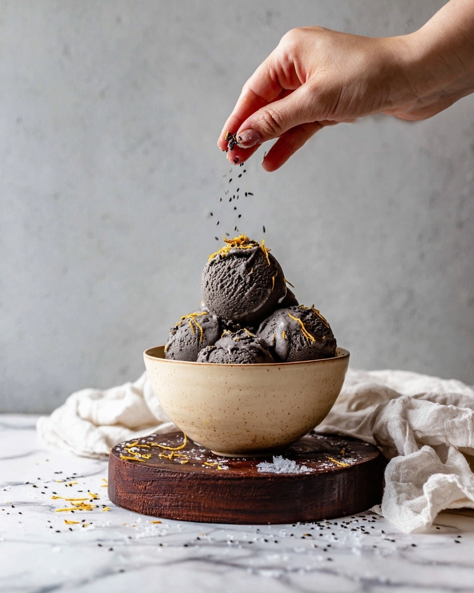 A bowl of black ice cream made up of five uneven scoops stacked together, each scoop showing a rough texture with small cracks and chips. Scattered thin yellow flakes are on top and around the scoops, some falling outside the bowl. A woman’s hand hovers above, sprinkling tiny black seeds over the top scoop. The ice cream sits inside a round beige bowl, which is placed on a dark brown round wooden plank. A white marbled surface underneath is strewn with more yellow flakes and black seeds. Next to the bowl on the right is a wrinkled off-white cloth with some flakes on it. The background is a plain grey wall. photo taken with an iphone --ar 4:5 --v 7