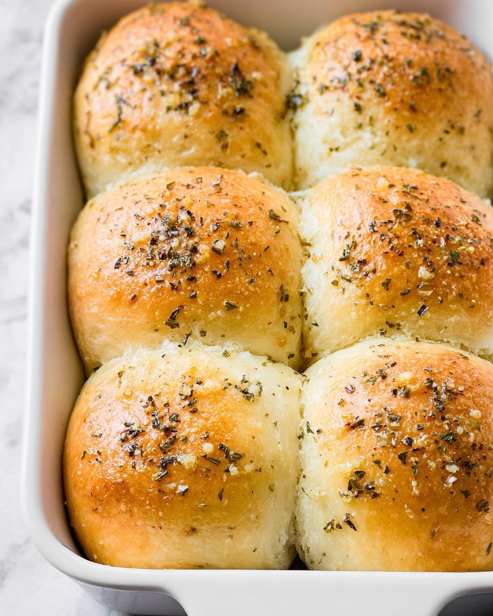 The image shows six soft, fluffy bread rolls tightly packed in a white baking dish. Each roll has a golden-brown top with a slightly shiny texture, sprinkled evenly with dried herbs and small bits of garlic or seasoning. The tops have a smooth but slightly uneven surface, and the rolls are touching each other, highlighting their pillowy and light appearance. The dish is placed on a white marbled surface, adding a clean and simple background to the warm, freshly baked bread. photo taken with an iphone --ar 4:5 --v 7