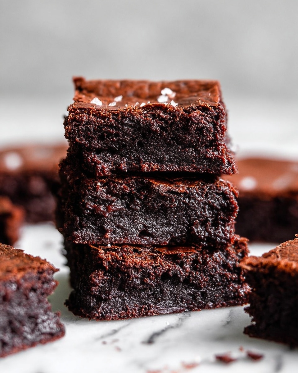 Three thick, dark brown brownies are stacked on top of each other in the center of the image, showing a rich, moist, and dense texture with a slightly crumbly surface. The top brownie has a few small bits of sea salt sprinkled on it. Around the stack, several more brownies are scattered on a white marbled surface, creating a casual, inviting look. The background is soft and blurred, keeping the focus on the brownies. photo taken with an iphone --ar 4:5 --v 7
