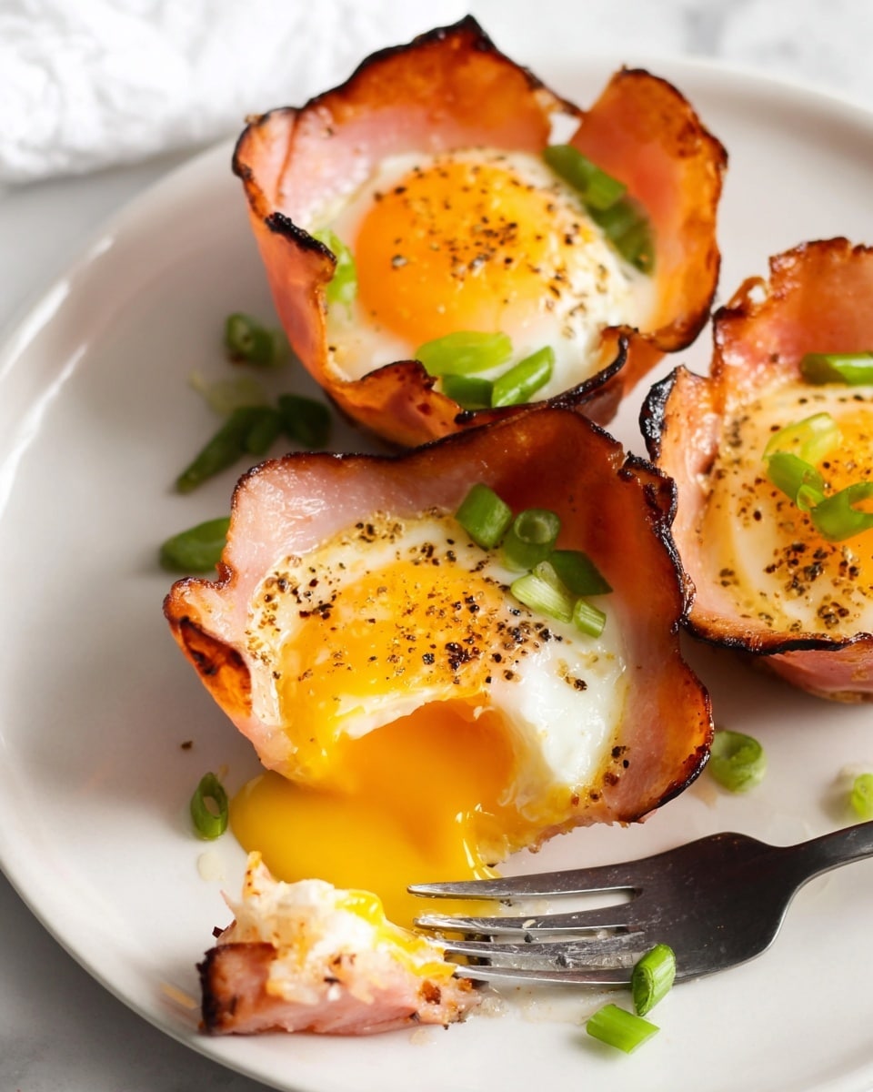The image shows three baked egg cups on a white plate, each made with a crispy, browned-edged slice of ham forming a cup shape. Inside each ham cup is a cooked egg with a bright yellow yolk and firm white, sprinkled with small black pepper flakes. Bright green chopped scallions are scattered on top of the eggs and around the plate. One egg cup is broken open with a piece of yolk and white on a fork resting on the plate. The plate is set on a white marbled surface. photo taken with an iphone --ar 4:5 --v 7