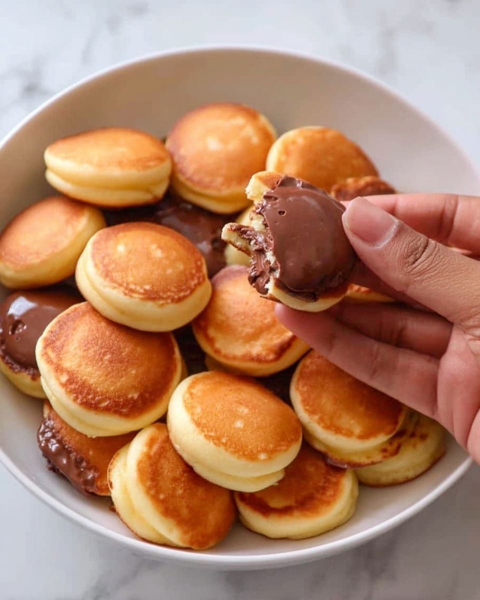 A white bowl filled with many small, round, golden-brown mini pancakes, each with a smooth, slightly crispy surface. Some pancakes are plain, showing their soft and fluffy texture, while a few have a layer of glossy chocolate spread on top, adding a rich dark brown contrast. A woman's hand is holding one mini pancake covered in chocolate, showing the thickness of the spread and the light interior of the mini pancake. The bowl is placed on a white marbled surface. photo taken with an iphone --ar 4:5 --v 7