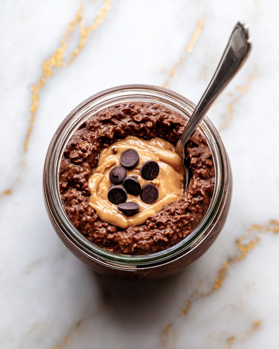 A small glass jar filled with a thick, dark brown chocolate oatmeal mixture, textured and slightly bumpy, topped in the center with a swirl of light brown peanut butter and six dark chocolate chips arranged in a small circle. A silver spoon rests inside the jar on the right side. The jar sits on a white marbled surface with gold vein patterns. photo taken with an iphone --ar 4:5 --v 7