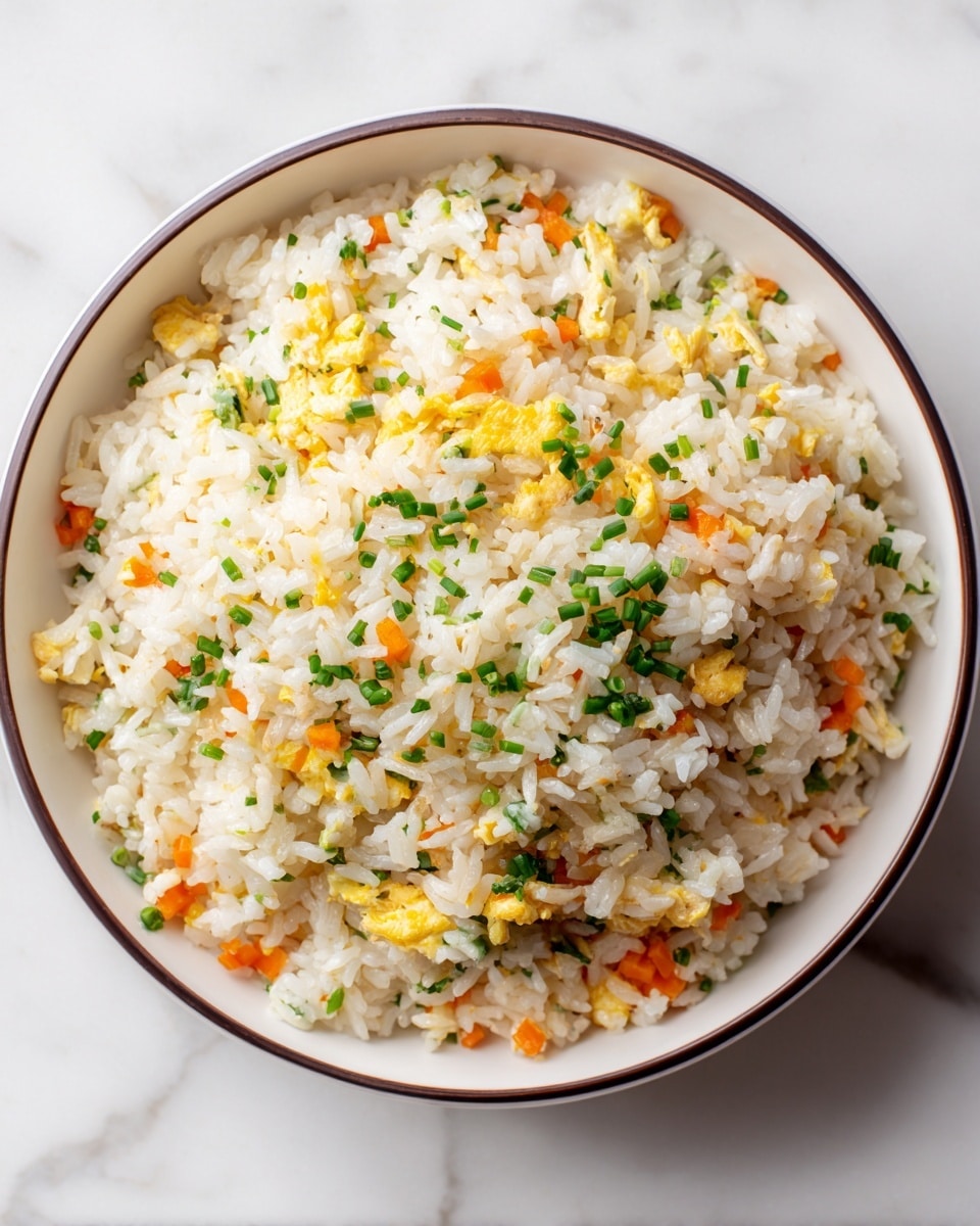 A close-up of a bowl filled with fried rice, showing one layer of rice grains mixed with small scrambled egg pieces and bits of orange carrot. The rice is white and slightly yellow from the egg, with green chopped chives sprinkled on top. The bowl is white with a thin dark rim, sitting on a white marbled surface. The texture looks soft and fluffy with small pieces of vegetables and egg spread evenly. Photo taken with an iphone --ar 4:5 --v 7