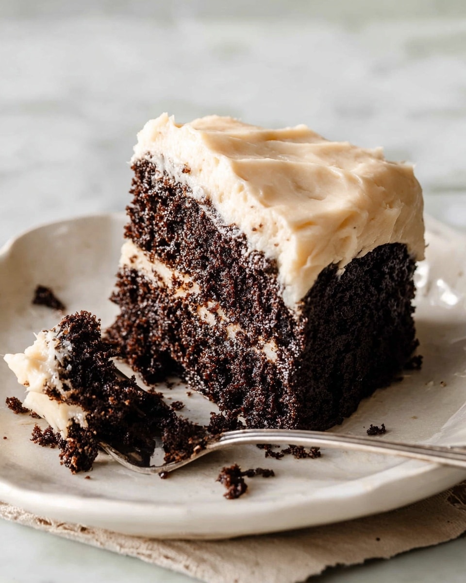 A single slice of dark chocolate cake with a thick, pale cream frosting layer on top sits on a white plate with a slightly wavy edge. The cake slice is cut in half with the front piece lying flat and the back piece standing upright, showing its moist, crumbly texture. A silver fork rests beside the cake, with some cake crumbs on it and scattered around the plate. The background has a soft white marbled texture. photo taken with an iphone --ar 4:5 --v 7