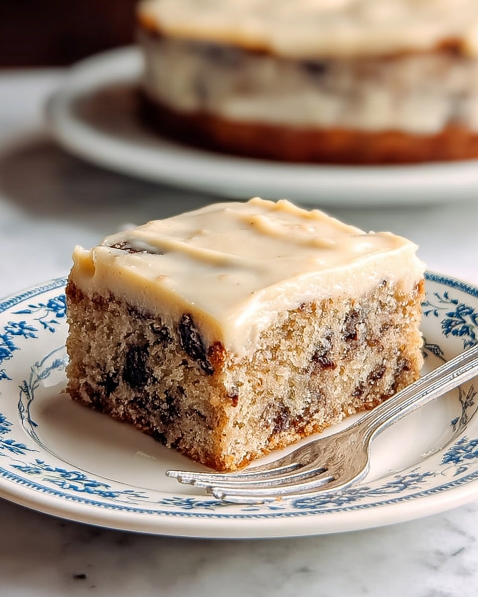 A single square piece of cake sits on a white plate with blue floral patterns along the rim. The cake has two clear layers: the bottom layer is dense with a light brown color and dark chocolate chips mixed throughout, while the top layer features a smooth, creamy beige frosting that slightly drapes over the edges. The cake appears moist with a crumbly texture on the sides. A silver fork rests on the right side of the plate. In the background, a larger round cake is visible, out of focus, on a white plate placed on a white marbled texture surface. photo taken with an iphone --ar 4:5 --v 7