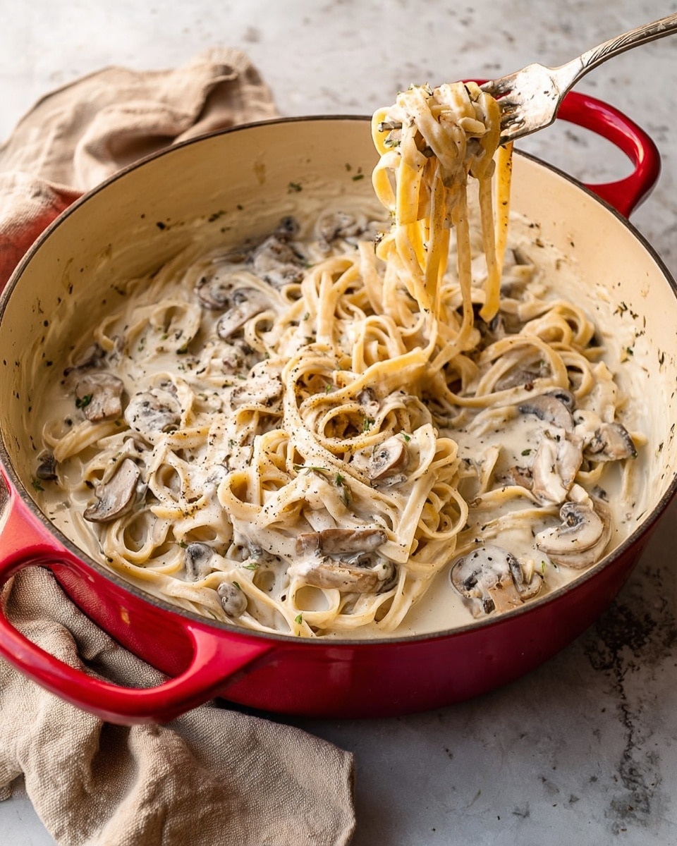 A red pot with a cream-colored inside holds creamy fettuccine pasta mixed with sliced mushrooms in a thick white sauce. The pasta strands are coated evenly, curling and layering over each other, with forks lifting a clump from the center. You can see specks of herbs and pepper sprinkled throughout the sauce, which has a smooth and rich texture. The pot sits on a white marbled surface, with a beige cloth casually placed nearby. photo taken with an iphone --ar 4:5 --v 7