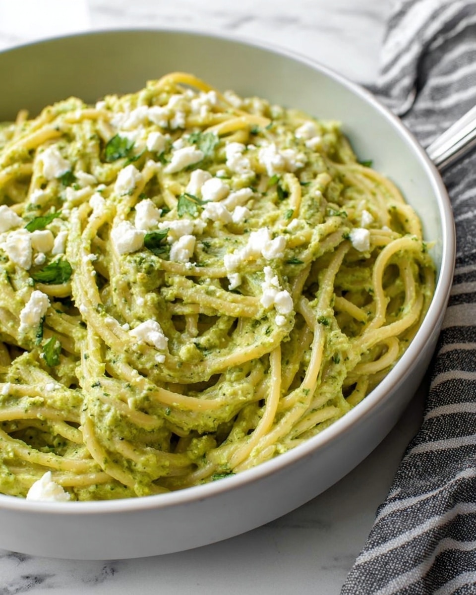 A close-up view of a white bowl filled with spaghetti coated in a thick, creamy green sauce, with some chopped green herbs mixed in. The noodles are covered and slightly mixed with the creamy sauce, creating a textured, smooth layer underneath that peeks through. Small white cheese crumbles are scattered evenly on top, adding contrast to the green pasta. The bowl sits on a white marbled surface with part of a gray and white striped cloth visible in the background. photo taken with an iphone --ar 4:5 --v 7