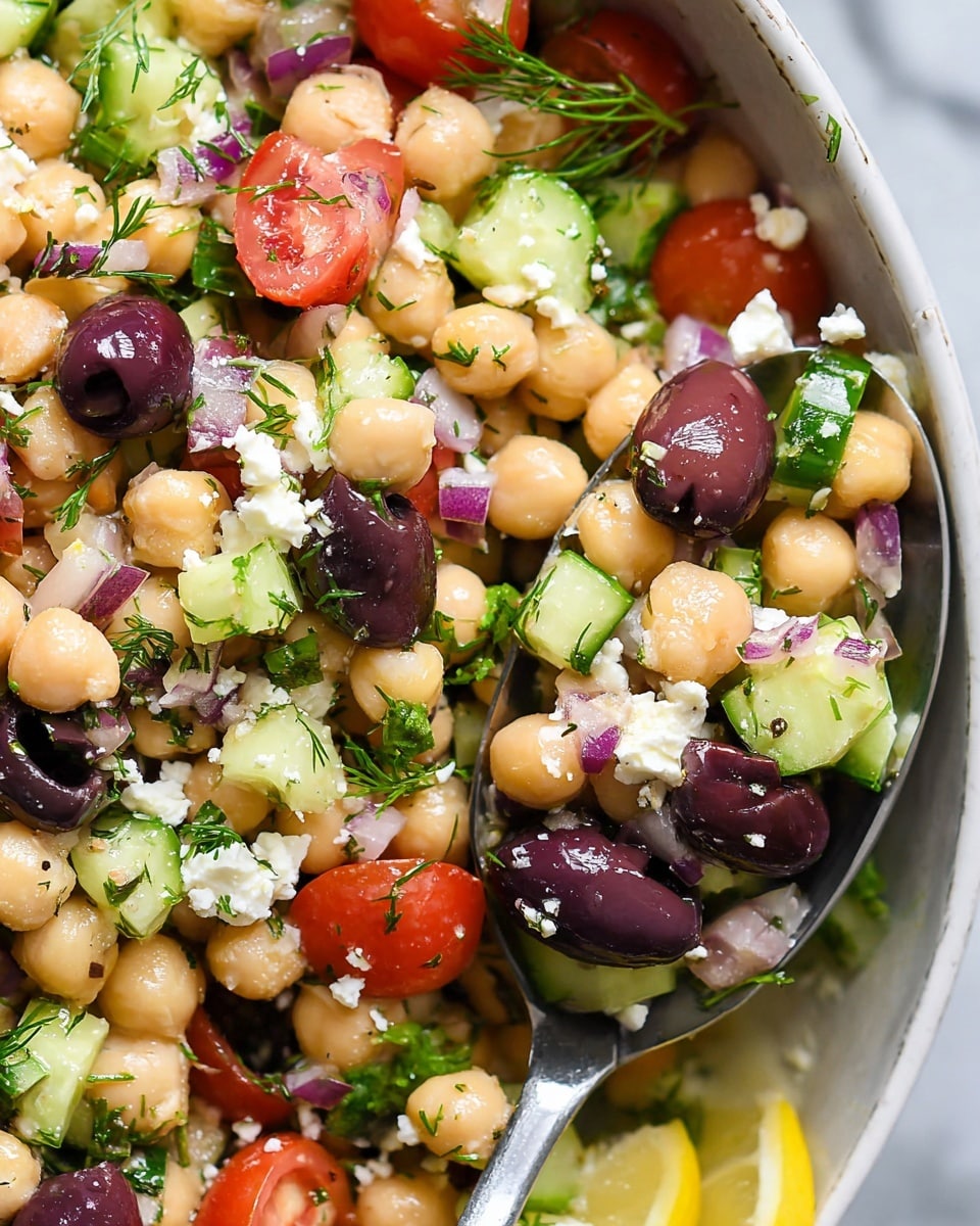 A close-up view of a fresh salad showing layers of beige chickpeas mixed with green cucumber chunks and halved bright red cherry tomatoes, along with dark purple Kalamata olives. Scattered throughout are small white crumbles of feta cheese and bits of chopped red onion adding more purple tones. Fresh green herbs like dill and parsley leaves are spread across, adding a touch of leafy texture. A silver spoon scoop is seen towards the right side, lifting a mix of these ingredients against a white marbled background, with a wedge of yellow lemon partly visible at the edge. Photo taken with an iphone --ar 4:5 --v 7