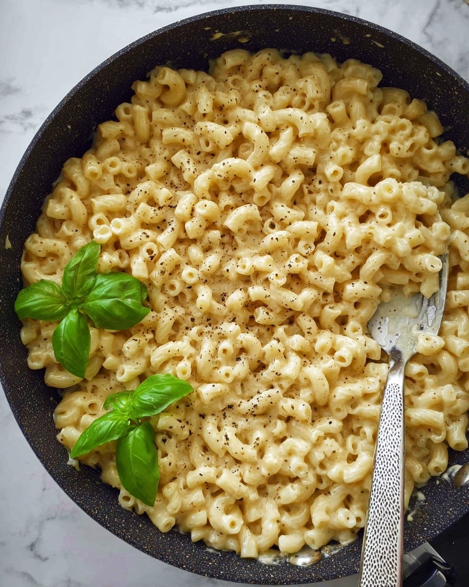 The image shows a single layer of creamy macaroni pasta in a large black pan. The pasta is light yellow, coated in a smooth cheese sauce with a slightly glossy texture. Black pepper is sprinkled evenly over the top, adding small dark specks. Three fresh green basil leaves are placed on one side, contrasting with the pale pasta. A silver spoon with a patterned handle is partially inside the pan, scooping up some pasta. The pan sits on a white marbled surface. photo taken with an iphone --ar 4:5 --v 7